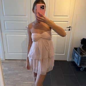 Ljusrosa axelbandslös klänning - Säljer en söt ljusrosa axelbandslös klänning med volanger. Klänningen har en kort längd och en knuten detalj framtill. Perfekt för sommarens varma dagar eller en speciell tillställning. 💖