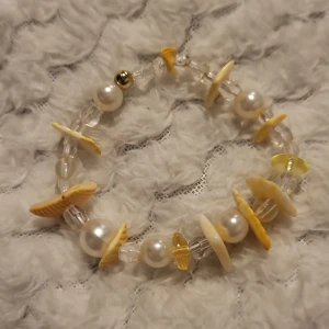 Armband med pärlor och snäckor - Charmigt armband med vita pärlor och snäckor i olika nyanser av gult och beige. Armbandet har även klara kristalliknande pärlor som ger en glittrig effekt. Perfekt för att ge en strandig känsla till din outfit.