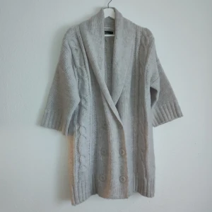 Beige stickad kofta från Gina Tricot - Mysig beige stickad kofta från Gina Tricot med kabelstickat mönster och stora knappar framtill. Koftan har trekvartsärmar och en bred krage som ger en avslappnad look. Perfekt för kyliga dagar!