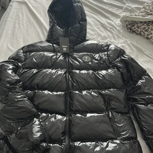 Svart pufferjacka från Trapstar - Säljer en svart pufferjacka från Trapstar med glansigt material och huva. Jackan har en dragkedja framtill och ett märke på bröstet. Perfekt för kalla dagar med sin quiltade design.
