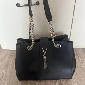 Svart axelväska från Valentino - Snygg svart axelväska från Valentino med silverkedjor och dekorativ tofsdetalj. Väskan har flera fack inuti och en dragkedja för säker förvaring. Perfekt för att bära med sig det viktigaste med stil.