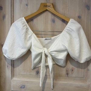 Vit knytblus från Zara - Säljer en söt vit blus från Zara med knytning framtill. Blusen har korta puffärmar och en lätt textur i tyget. Perfekt för varmare dagar och ger en romantisk touch till din outfit.