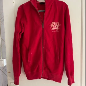 Röd huvtröja med dragkedja - Säljer en röd huvtröja med dragkedja framtill. Tröjan har ett tryck med texten 'Peace & Love' på bröstet och ryggen, samt datumet 28 juni - 2 juli 2011. Den har långa ärmar och en bekväm passform. Perfekt för höst och vår!