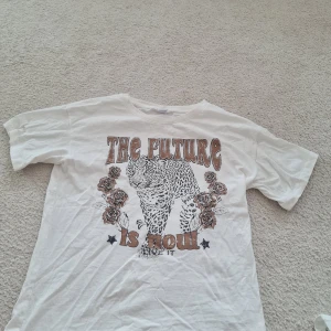 Vit t-shirt med leopardtryck från Lindex - Säljer en cool vit t-shirt från Lindex med ett stort leopardtryck och texten 'The Future is Now' på framsidan. T-shirten har korta ärmar och är i storlek 170, vilket passar ungdomar. Perfekt för en avslappnad och trendig look!