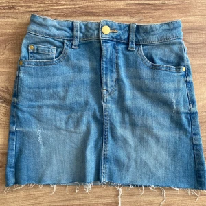 Jeanskjol med rå kant - Säljer en snygg jeanskjol med rå kant. Kjolen har en klassisk blå jeansfärg och är i en kort modell. Den har fem fickor och stängs med en knapp och dragkedja framtill. Perfekt för en avslappnad och cool look!