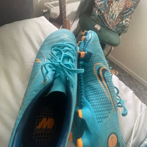 Blå fotbollsskor från Nike - Säljer ett par blå fotbollsskor från Nike. Skorna har en snygg design med orange detaljer och snörning. De är i bra skick och redo för nästa match!