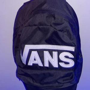 Svart ryggsäck från Vans - Säljer en svart ryggsäck från Vans. Den har ett stort fack med dragkedja och ett mindre fack på framsidan. Insidan har ett schackrutigt mönster och elastiska fickor. Väskan är i bra skick och perfekt för skolan eller vardagsbruk.