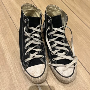 Svarta Converse All Star sneakers - Säljer ett par svarta Converse All Star sneakers i storlek 39. Skorna är välanvända med vita snören och en vit sula som har blivit lite smutsig. De har den klassiska höga modellen med snörning och Converse-loggan på sidan. Perfekta för en casual look!