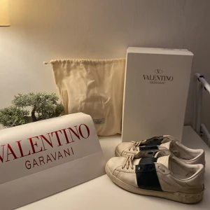 Valentino Garavani sneakers - Säljer ett par snygga och stilrena Valentino Garavani sneakers i vitt med en mörkblå rem över snörningen. Skorna är i skinn och har en klassisk design som passar till många olika outfits. De är i storlek herr 45 och kommer med originalförpackning och dustbag.