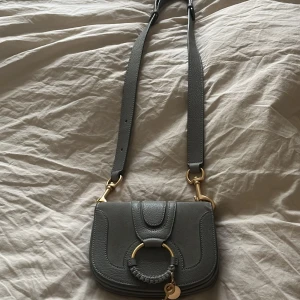 Grå axelväska från See By Chloé - Säljer en superfin grå axelväska från See By Chloé i skinn och mocka. Den har snygga gulddetaljer och en flätad ring framtill med en liten nyckel och logotyp. Perfekt för både vardag och fest! 👜✨Nästan aldrig använd, priset kan diskuteras