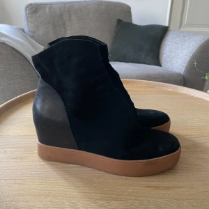 Svarta kilklackade boots - Snygga svarta boots med kilklack och en brun sula. De är gjorda i mocka och har en stilren design som passar perfekt för höst och vår. Skorna har en tight passform och är i mycket bra skick. Perfekta för en casual look eller en utekväll!