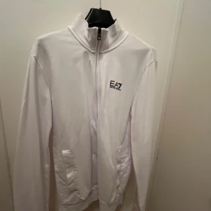 Vit tröja från Emporio Armani, EA7 - Säljer en stilren vit tröja från Emporio Armani med dragkedja och hög krage. Tröjan har en diskret logga på bröstet och svarta ränder på ryggen. Perfekt för en casual look eller träning. Den är i mycket bra skick och passar till alla säsonger. Strl M.
