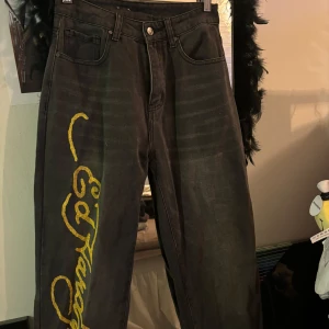 Svarta jeans med gul text från Ed Hardy - WOMENS BORN-WILD BOYFRIEND JEAN JEANS” - Ed Hardy.  Coola svarta jeans från Ed Hardy med gul text 'Born Free'och stort tryck längs benet. De har en loose fit och är perfekta för en avslappnad stil. Jeansen har hög midja men går att ha som mid rise för en mer avslappnad stil. Köpta för 1500 men säljer gör 800, köpta men aldrig använda endast testade.  Mid rise/högmidjade beroende på hur man vill ha dom! Snygg baggy fit, jag