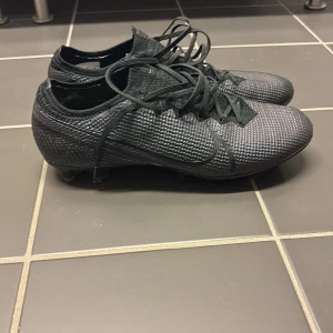 Svarta fotbollsskor från Nike - Säljer ett par svarta Nike fotbollsskor i mycket bra skick. Skorna har en tight passform och är perfekta för fotbollsträning. De har snörning och en stilren design med NikeGrip-teknologi för extra grepp. Perfekta för dig som vill ha stil och funktion på planen!                    Vapor 13 AG, knappt använda!!!!                  Nypris: svåra att få tag på idag, runt 3000kr