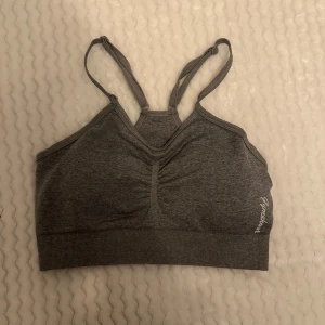 Grå sport-bh från Gymshark - Säljer en grå sport-bh från Gymshark i mycket bra skick. Den har en snygg och enkel design med justerbara axelband och en liten logga på sidan. Perfekt för träning eller vardagsbruk. Materialet är mjukt och stretchigt för bästa komfort.