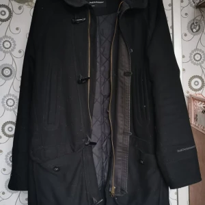 Svart duffel från Peak Performance - Säljer en stilren svart duffel-kappa från Peak Performance i ullblandning. Den har en quiltad insida och stängs med dragkedja och knappar. Nypris 3000kr. Perfekt för höst och vinter med sin långa design och huva. Passar både till vardags och lite finare tillfällen. Se bild för skicka. 