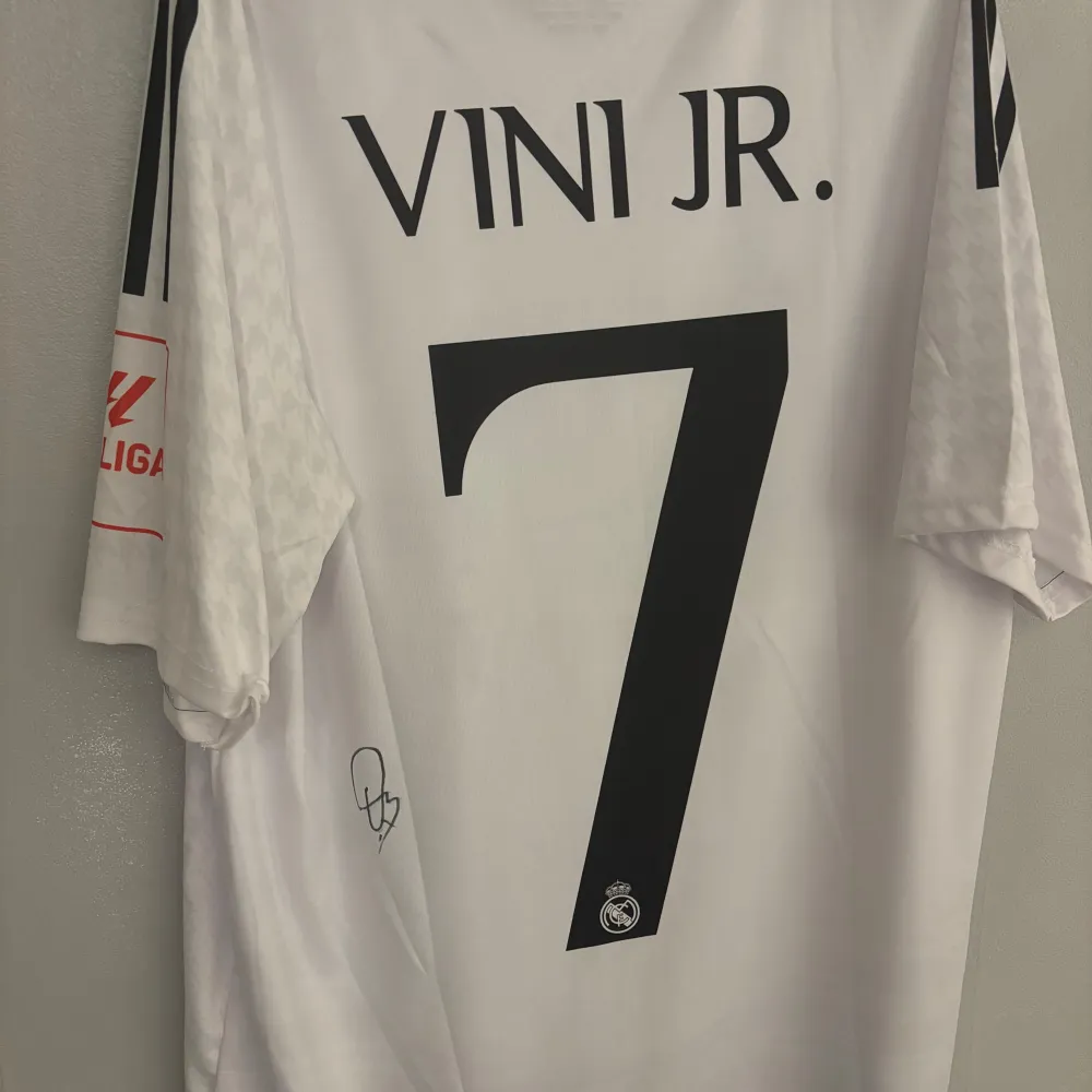 Säljer en vit Real Madrid fotbollströja i mycket bra skick. Tröjan har korta ärmar och är prydd med klubbens emblem och sponsorlogga. Den har även ett stort nummer 7 på ryggen. Perfekt för alla fans av laget!. T-shirts.