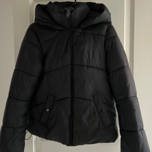 Svart pufferjacka från Vero Moda - Säljer en svart pufferjacka från Vero Moda i storlek XS. Jackan har en dragkedja framtill och praktiska fickor med knappar. Perfekt för kalla dagar med sin varma och vadderade design. Huvan ger extra skydd mot väder och vind. Passar perfekt till både vardag och utflykter!