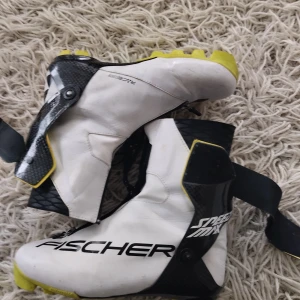 Vita skidpjäxor från Fischer - Säljer ett par vita längdskidpjäxor från Fischer i bra skick. De har en cool design med svarta och gula detaljer samt snörning och dragkedja för bra passform. Perfekta för vinterns äventyr i skidbacken! 