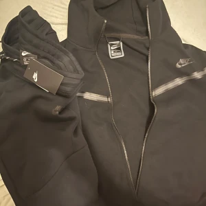 Svart hoodie från Nike - Säljer en svart hoodie från Nike i nytt skick. Den har en dragkedja framtill och en klassisk Nike-logga på bröstet. Perfekt för både träning och vardag. Huvan är justerbar med snören och materialet känns mjukt och bekvämt. Passar perfekt till höst och vinter.