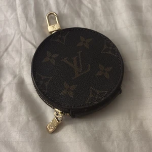 Rund söt plånbok  - Säljer en superfin rund plånbok i klassiskt monogrammönster. Den är i mörkbrunt skinn med guldfärgade detaljer och dragkedja. Perfekt för att hålla småsaker eller mynt. Den har en praktisk karbinhake för att fästa i väskan. 👜✨