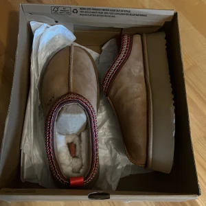  UGG Tazz - Säljer ett par bruna mockaskor från UGG i nytt skick. De har en mysig foder av päls och en snygg röd och svart broderad kant. Perfekta för höst och vinter med en bekväm passform och robust sula. Skorna är i storlek dam 40