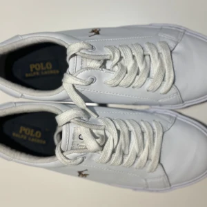 Vita sneakers från Polo Ralph Lauren - Säljer ett par vita sneakers från Polo Ralph Lauren i jätte bra skick. Endast använd en gång. De har en klassisk design med snörning och den ikoniska Polo-loggan på sidan. Perfekta för en avslappnad stil och passar till det mesta. Skorna är bekväma och stilrena, idealiska för både vardag och fest.