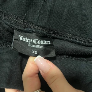 Svarta byxor från Juicy Couture - Säljer ett par svarta byxor från Juicy Couture i storlek XS. Byxorna har en mjuk och glansig finish med fickor och snygga knappar. Perfekta för en avslappnad men stilren look. 🖤knapparna är lite rostiga men det syns inte, har knappt andvänt dom💕