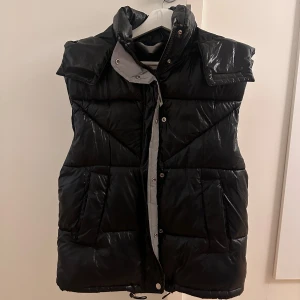 Svart dunväst med huva - Säljer en svart glansig dunväst i mycket bra skick. Västen har en praktisk huva och stängs med både dragkedja och knappar. Den har en reflex längst med dragkedjan.