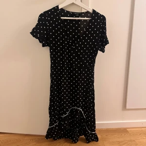 Svart prickig klänning från Monki - Säljer en superfin svart klänning från Monki med vita prickar. Den har en omlott-design , korta ärmar och Volangdetaljer längst ner. Klänningen är i mycket bra skick och passar perfekt till vår och sommar!