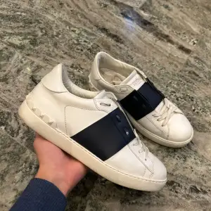 Säljer ett par snygga Valentino Garavani sneakers i vitt skinn med en cool blå detalj över snörningen. Skorna är i bra skick och har en stilren design med de klassiska Valentino-nitarna på hälen. Perfekta för både vardag och fest!