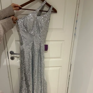 Glittrig silverklänning - Säljer en fantastisk glittrig silverklänning med oneshoulder-design. Perfekt för nyårsfesten eller en glamorös kväll. Klänningen är lång och har en elegant passform som verkligen fångar ljuset. Den är i mycket bra skick och redo att bäras av någon som vill glänsa!