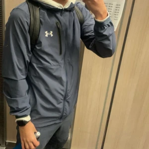 Blå trackasuit från Under Armour - Säljer en blå vindjacka och byxor från Under Armour i mycket bra skick. Jackan har en dragkedja framtill och är perfekt för träning eller vardagsbruk. Den är långärmad och har en sportig look. Perfekt för höst och vår! Nypris 1700kr