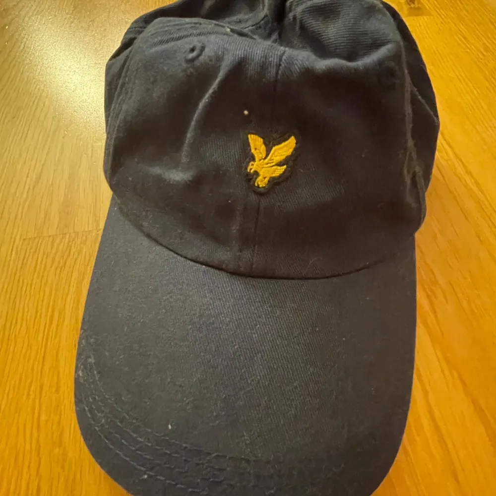 Snygg svart keps från Lyle & Scott med deras ikoniska gula logga broderad framtill. Kepsen är i bomull och har en justerbar rem baktill för perfekt passform. Perfekt för både vardag och sportiga aktiviteter.. Accessoarer.