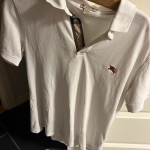 Vit pikétröja från Burberry - Säljer en klassisk vit pikétröja från Burberry i bra skick. Den har en broderad röd logga på bröstet och stiliga knappar med Burberrys ikoniska rutiga mönster på insidan av knappslån. Perfekt för en stilren och avslappnad look!