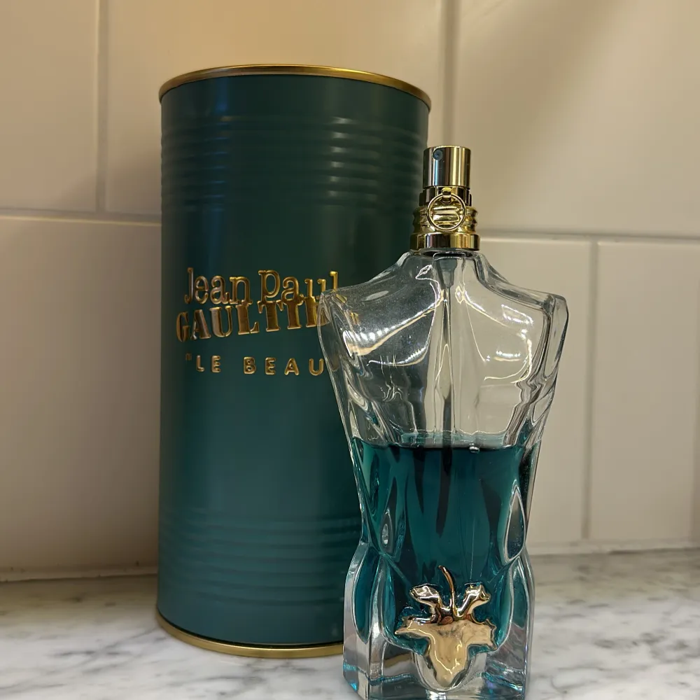 Säljer en Jean Paul Gaultier Le Beau Eau de Toilette. Flaskan är formad som en manlig torso i glas med en blågrön vätska inuti och en guldig detalj framtill. Den kommer i en elegant grön metallcylinder med guldtext. Perfekt för den som vill ha en unik och stilren parfymflaska på hyllan!😇🌟. Parfym.