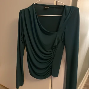 Grön långärmad topp från Gina Tricot - Snygg grön långärmad topp från Gina Tricot i ett mjukt och stretchigt material. Toppen har en draperad design framtill som ger en elegant look. Perfekt för både vardag och fest!