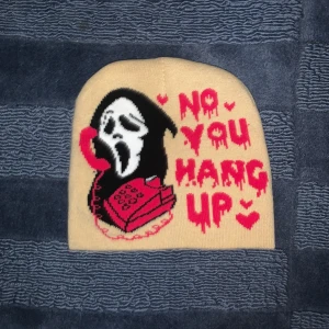 Beige mössa med tryck - Cool beige mössa med ett tryck av en skrikande figur i svart och vitt, som håller en röd telefon. Texten 'NO YOU HANG UP' i röd färg med små hjärtan runt. Perfekt för att ge en edgy touch till din outfit!