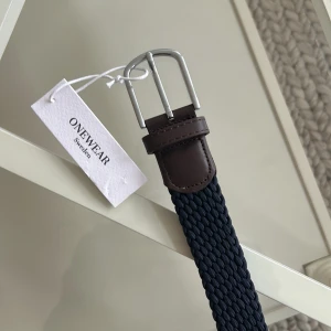 Mörkblått bälte från Onewear - Snyggt mörkblått bälte från Onewear Sweden med silverfärgat spänne och bruna skinn-detaljer. Perfekt för att ge en stilren touch till din outfit. Passar både till vardag och fest. Ett måste i garderoben för den modeintresserade!