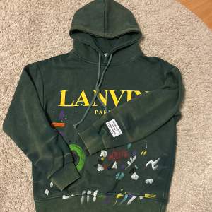 Säljer en grön hoodie från Lanvin med coola färgstänk och tryck framtill. Den har en stor huva och ribbade muddar. Perfekt för en avslappnad stil med en konstnärlig touch. Passar både till vardags och chillkvällar.