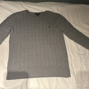 Grå stickad tröja från Ralph Lauren - Säljer en snygg grå stickad tröja från Ralph Lauren i storlek XL. Tröjan har ett klassiskt kabelstickat mönster och en liten broderad logga på bröstet. Perfekt för höst och vinter, både stilren och bekväm. Passar till både vardag och lite finare tillfällen. 