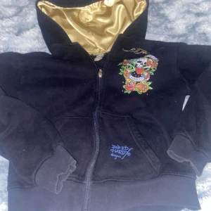Svart hoodie från Ed Hardy - svart hoodie från Ed Hardy med ett färgglatt tryck av rosor och en dödskalle. Texten 'Love Dies Hard' ger en edgy vibe. Insidan av huvan är i guldigt tyg som ger en lyxig känsla. Fråga alltid ol ni vill se den på så kan ja visa. Annars bra bara några fåtal diamanter som ramlat av fram o bak.