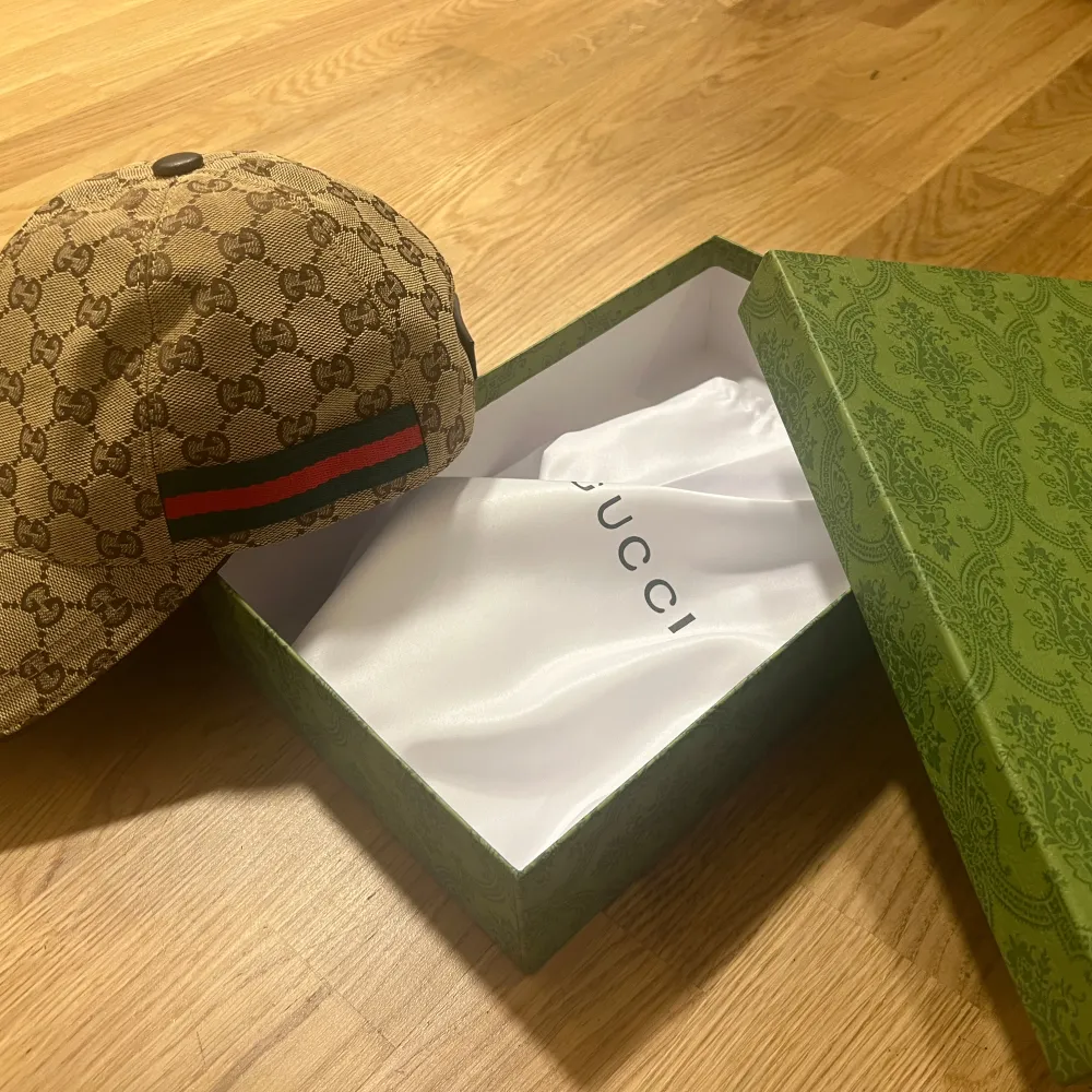 Säljer en snygg beige keps från Gucci med det klassiska GG-mönstret. Kepsen har en grön och röd randdetalj på sidan och kommer i sin originalförpackning. Perfekt för att ge din outfit en lyxig touch!. Accessoarer.