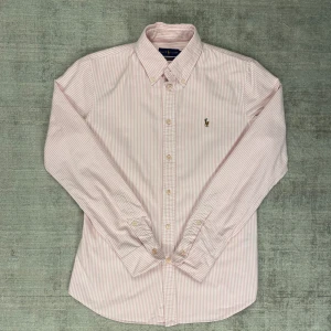 Rosa och vit randig skjorta från Ralph Lauren - Säljer en snygg rosa och vit randig skjorta från Ralph Lauren i storlek M. Skjortan är i bomull och har en klassisk krage med knappar. Den är långärmad och har en broderad logga på bröstet. Perfekt för både vardag och fest!