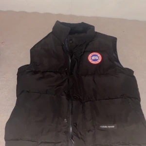 Svart dunväst från Canada Goose - Säljer en svart dunväst från Canada Goose i mycket bra skick. Västen har en broderad logga på bröstet och stängs med dragkedja och knappar. Perfekt för kyliga höst- och vinterdagar. Storleken är S, pris kan diskuteras 