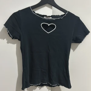 Svart topp med hjärtdetalj - Snygg svart topp från Bershka med en söt hjärtformad detalj vid halsen. Toppen har korta ärmar och en vågig kant vid ärmslut och nederkant. Perfekt för en casual look eller en utekväll. 🖤