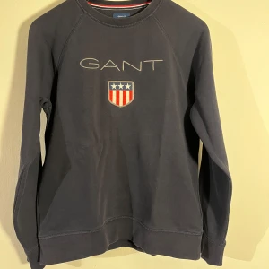 Mörkblå sweatshirt från GANT - Säljer en stilren mörkblå sweatshirt från GANT med broderad logga och emblem på bröstet. Tröjan har långa ärmar och en klassisk rund halsringning. Perfekt för en avslappnad stil.