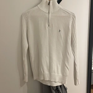 Vit stickad tröja från Peak Performance - Säljer en stilren vit stickad tröja från Peak Performance. Tröjan har en dragkedja vid halsen och ett diskret logotypbroderi på bröstet. Perfekt för kyligare dagar med sin ribbade krage och ärmslut.
