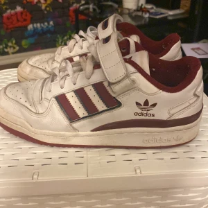 Vita och vinröda sneakers från Adidas - Säljer ett par klassiska vita sneakers från Adidas med vinröda detaljer och kardborreband. Skorna har de ikoniska tre ränderna på sidan och Adidas-loggan på hälen. Perfekta för en retro look! Pris ej hugget i 🪨, tvättar även skorna innan frakt 