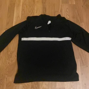 Svart långärmad tröja från Nike - Säljer en svart långärmad tröja från Nike med en vit horisontell rand över bröstet och en liten dragkedja vid halsen. Perfekt för träning eller vardagsbruk. Tröjan har en sportig look med en diskret Nike-logga på bröstet.